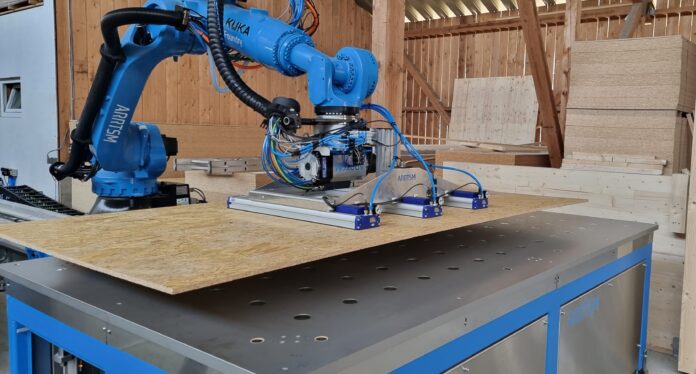 A robot uses a specialized vacuum attachment to lift panel board material from the inventory stack to the cutting table in preparation for fabricating a wall panel for a residential project.